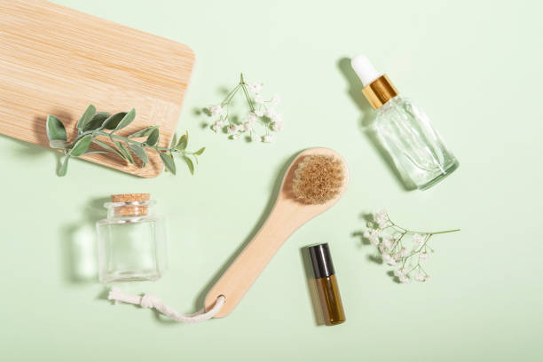 Natural wooden massage brush and glass cosmetic bottles on light green background with white flowers. Body and face treatment and spa. natural beauty products. massage oil. Copy space. Flat lay